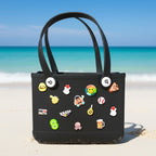 Mango Muffins black PVC tote bag decorated with fun charms, set on a sandy beach with the ocean in the background.