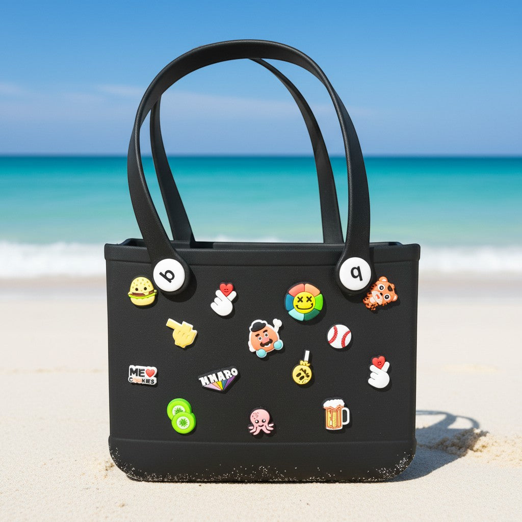 Mango Muffins black PVC tote bag decorated with fun charms, set on a sandy beach with the ocean in the background.