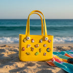 Mango Muffins yellow waterproof PVC tote bag with colorful charms, on a beach with ocean background.